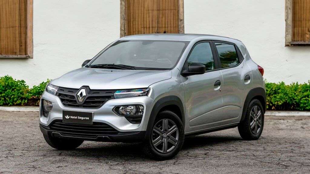 A imagem mostra um carro Renault Kwid de cor prata estacionado em uma superfície pavimentada. O veículo está posicionado de frente, exibindo a grade frontal com o logotipo da Renault no centro. Os faróis dianteiros são visíveis, assim como os espelhos retrovisores laterais e as rodas. Ao fundo, há uma parede branca com duas janelas de madeira fechadas e uma fileira de plantas verdes na base da parede. A placa do carro exibe o texto "Natal Seguros". Esta imagem ilustra o blog post sobre seguro kwid.